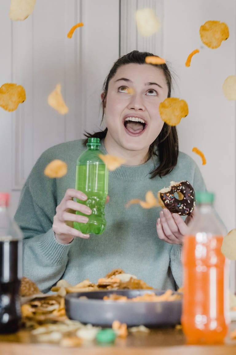 Cheerful young lady with opened mouth holding glazed chocolate bitten donut and bright bottle with fresh drink and surprising at shower of crisps and chips
