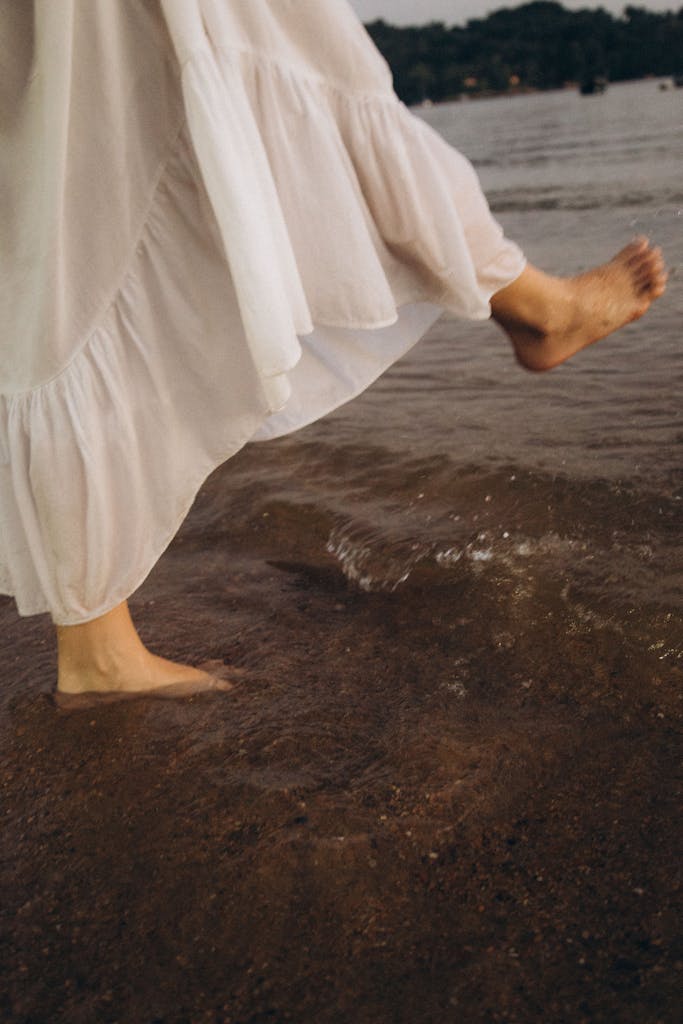 Close-up of a woman walking barefoot in water with a white flowing dress.