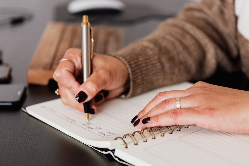Crop unrecognizable female with stylish manicure sitting at black desk with keyboard and smartphone and planning for healthy eating with silver pen in notepad