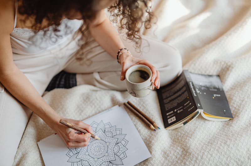 Woman colouring mandala while holding coffee on cozy blanket, during autumn, symbolizing rest and calm