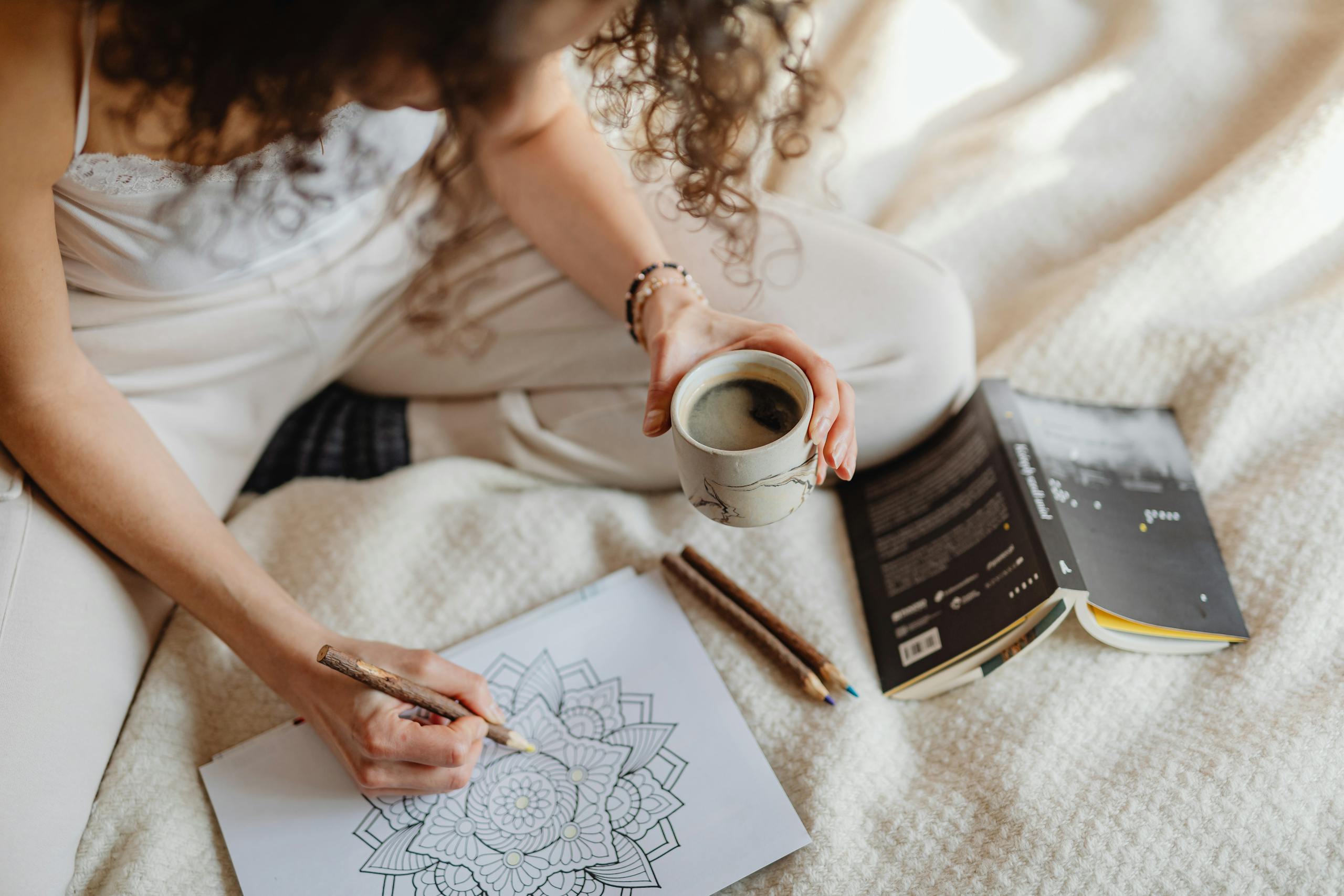 Woman colouring mandala while holding coffee on cozy blanket, during autumn, symbolizing rest and calm